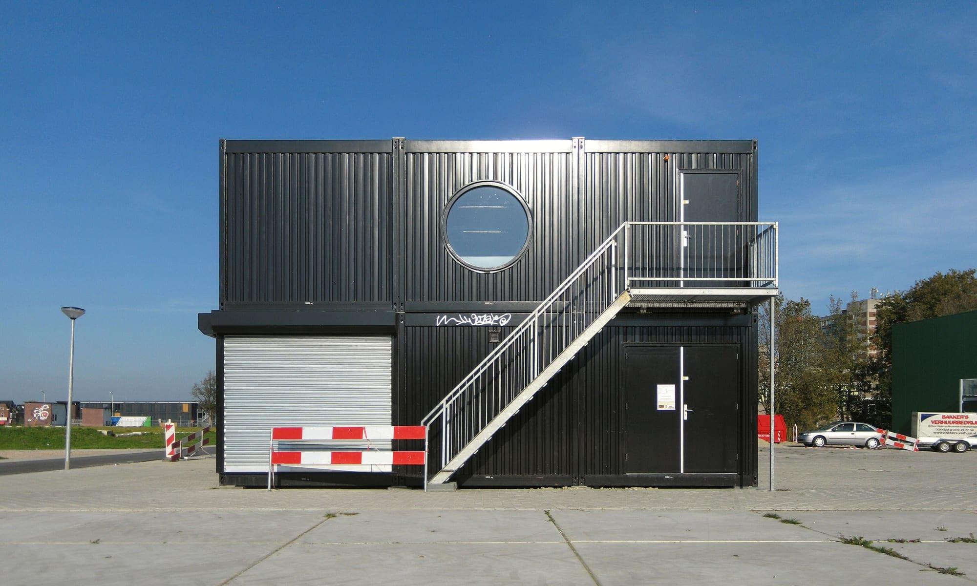 A two-storey black container building with a round window and an external steel staircase