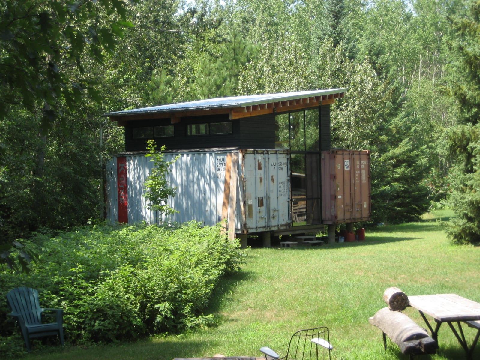 A small cabin made from two shipping containers under a raised roof, set in a green garden