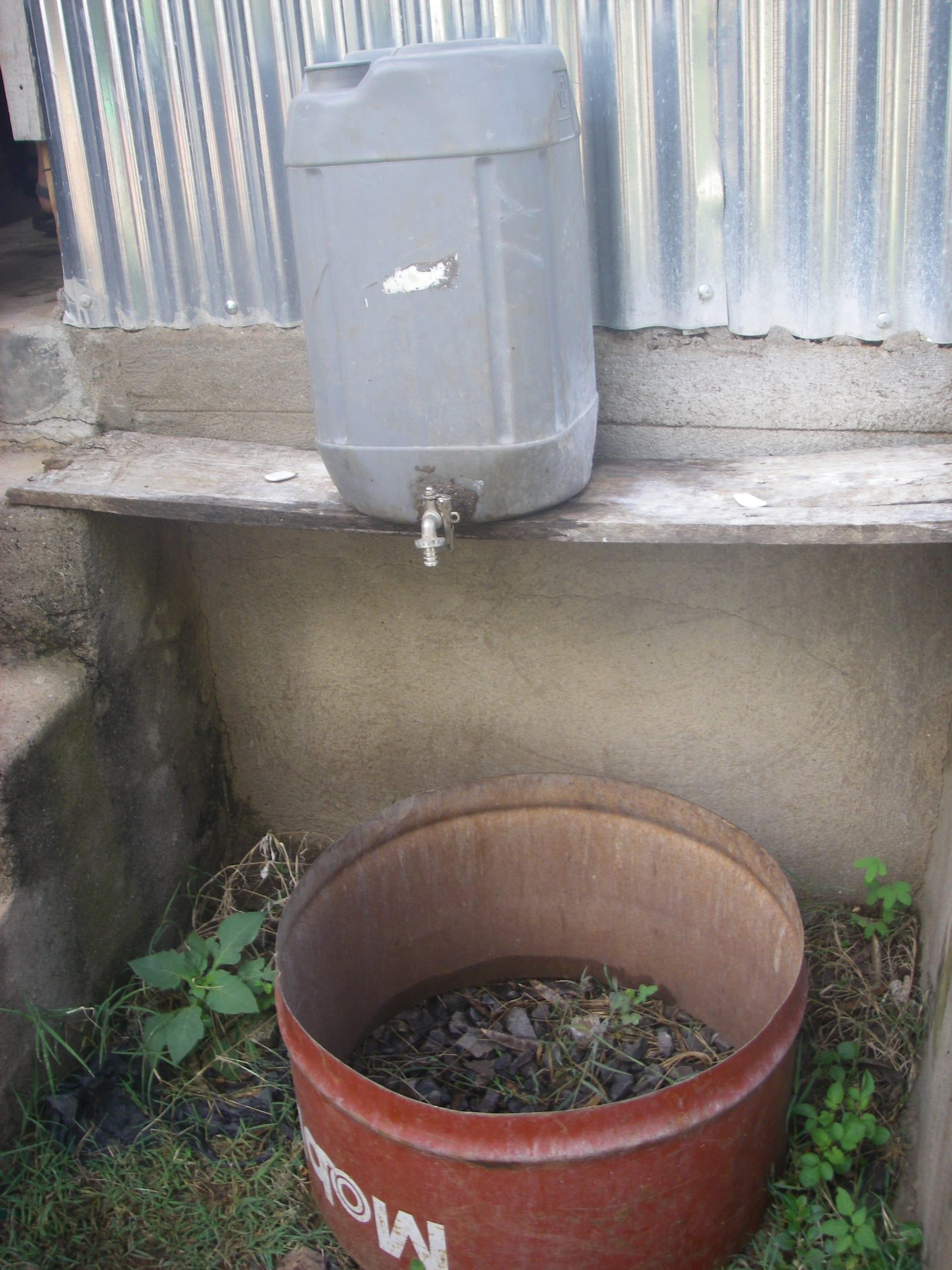 A jerrycan with a tap on a concrete ledge above a basin made from a cut-down steel drum