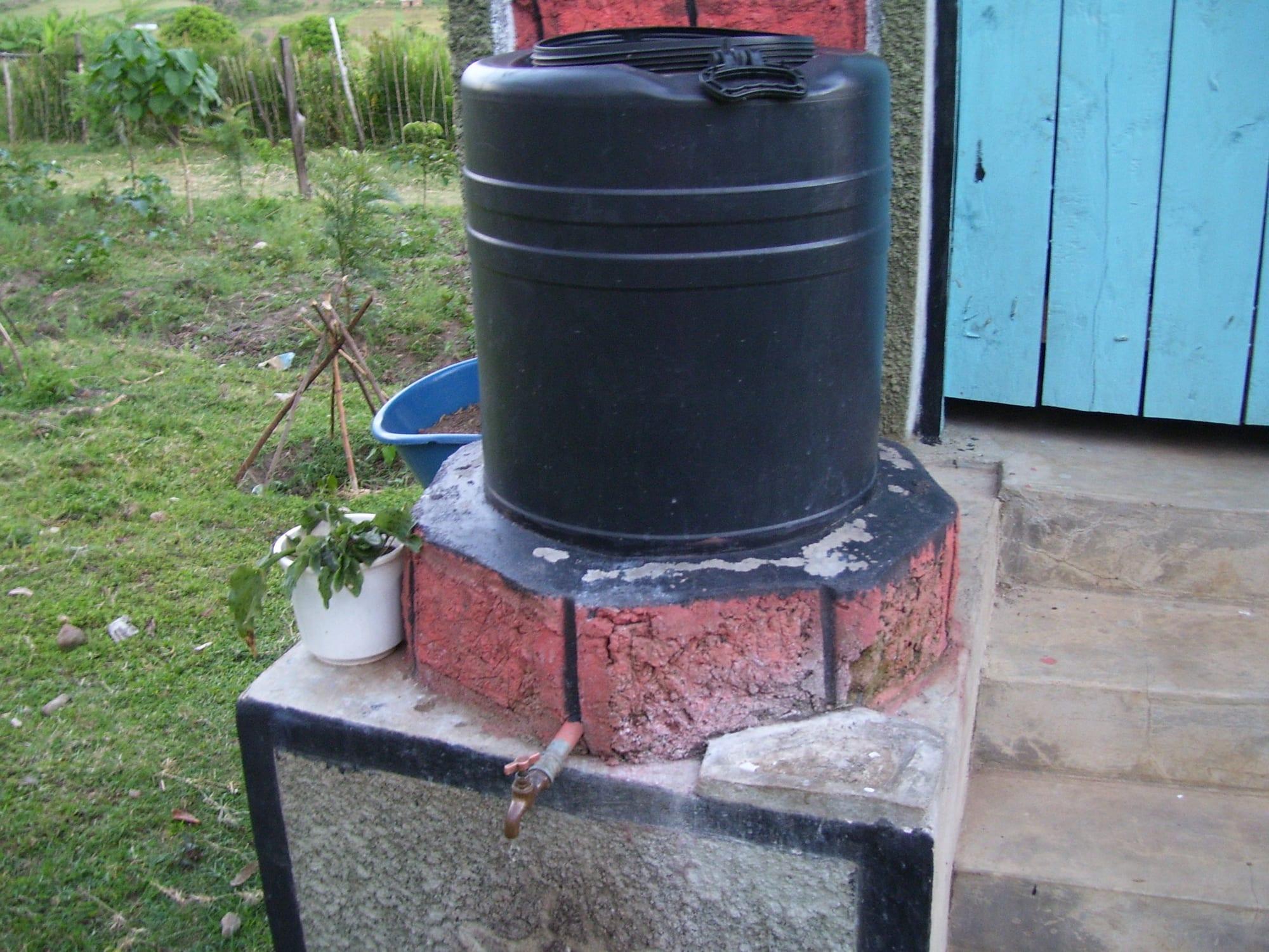 A black water tank on a round brick plinth with a tap, filled from a roof gutter, beside a toilet block
